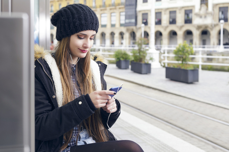 Young woman waiting on station platform, using smart phone sitting on a bench. Tourist texting message and planning route. Outdoors.の写真素材