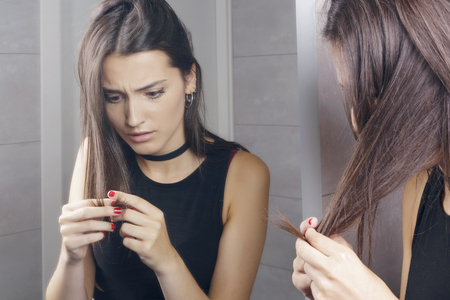 Surprised young woman finding damaged hair, standing in front of a mirror, indoors.の写真素材