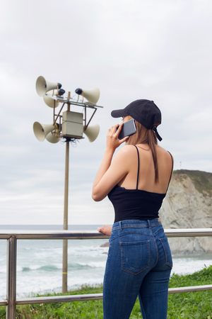 Young woman having phone conversation while standing near sea, outdoors near a speaker emergency post.の写真素材