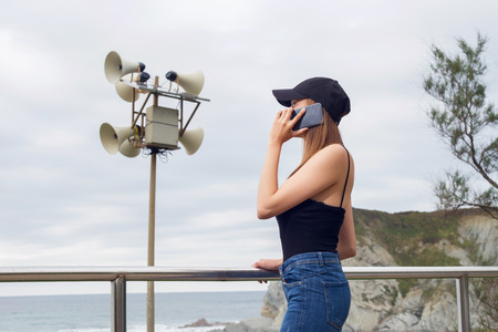 Young woman having phone conversation while standing near sea, outdoors near a speaker emergency post.の写真素材