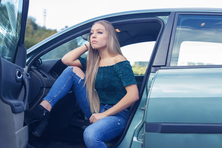 Pensive blonde woman in the car with opened door, outdoors, in a cloudy day.の写真素材