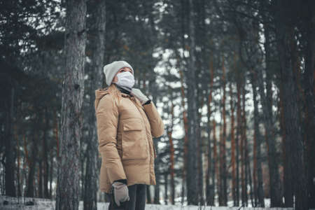 A young woman with blond hair in a medical mask walks in the winter forest.の写真素材