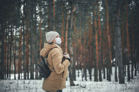 A young girl in winter clothes and in a medical mask is walking in the forest.