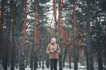 An attractive girl with blond hair and a cup of hot drink walks in the winter forest.の写真素材