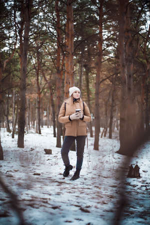 Attractive girl in winter clothes and and a cup of hot drink is walking in the forest.の写真素材