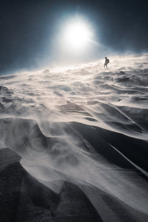 A lone hiker in warm clothes, in the cold winter walks on the frozen mountain, on sunny day with clear blue sky and strong windの写真素材