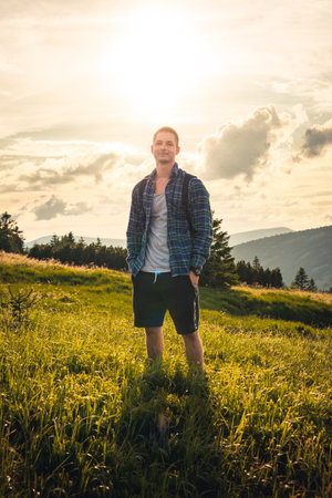 Young man with backpack standing in grass with sunset right behind him, Giant Mountains, Czech Republicの写真素材