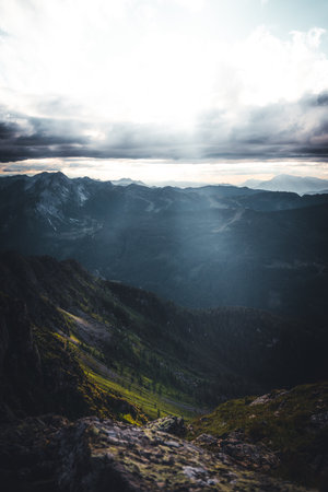 The brilliant sun is shining brightly through the fluffy clouds that are gracefully drifting over the majestic mountains in the distanceの写真素材