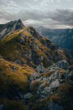 A picturesque mountain with a blanket of grass and scattered rocks adorning its surface, while a cloudy sky forms a dramatic backdrop aboveの写真素材