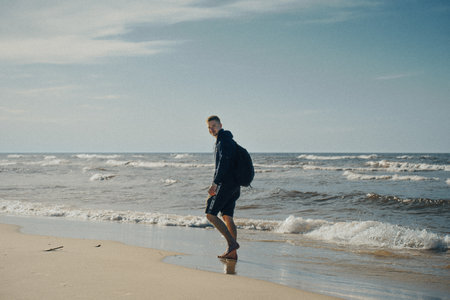 A man is leisurely walking along the picturesque beach while carrying a backpack on his back, enjoying the scenic view around himの写真素材