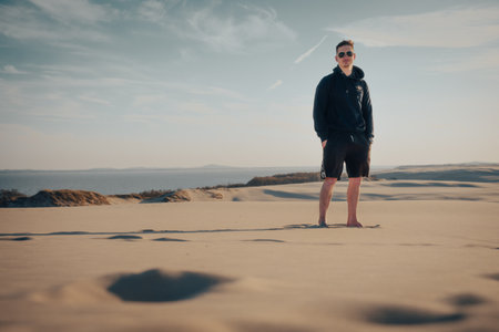 A man is standing atop a tall sand dune, wearing a hoodie and shorts, surrounded by the expansive landscape of the desertの写真素材