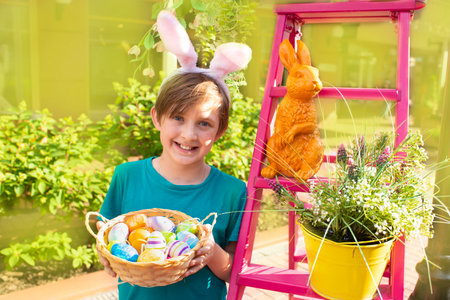 happy boy with bunny ears on his head and a basket of colorful easter eggs. a joyful child celebrates Easter, stands at the scenery for the holiday.の写真素材