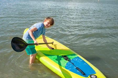 a happy boy enters the water, holds a SUP board with his hand, is going to ride on the board. 10 year old joyful child spends summer holidays doing active water sportsの写真素材