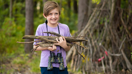 happy 10 year old boy on a hike, in a camp, holding a firewood. a joyful child holds branches for a fire, stands in front of a hut made of branches.の写真素材
