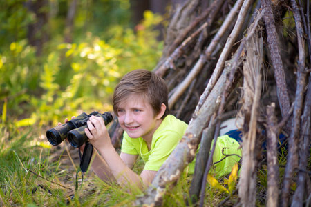 10 year old boy looks through binoculars, lies in a hut made of branches in a summer camp. a happy child spends his summer holidays hiking, outdoors, in the forest.の写真素材
