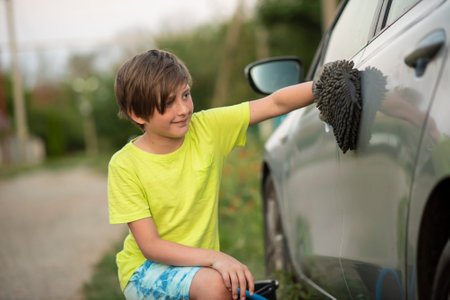 A 10-year-old boy helps his parents wash or dry the car. the child washes or wipes the car with a washcloth-mitten.の写真素材