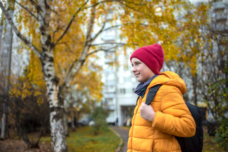 An 11-year-old child in a red hat, yellow jacket with a backpack goes to school in the fall, in cold weather. a smiling boy walks from home to study, past birch trees with orange fの写真素材