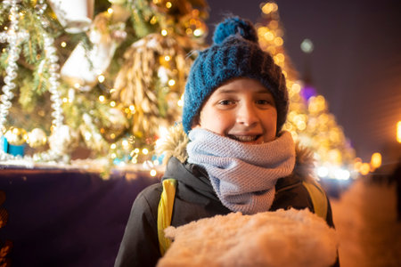a smiling boy in a winter hat and jacket holds a piece of ice and snow in his hands, stands in front of a decorated Christmas tree in garlands, at a New Year's fair in the city,の写真素材
