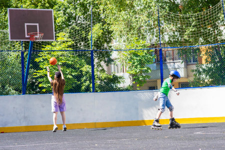 Two children on roller skates play basketball on the playground in summer