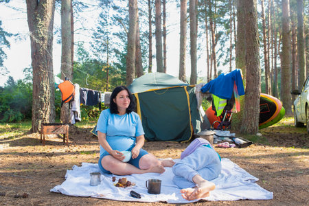 41 year old pregnant woman resting sitting on a blanket in front of a tourist tent, getting ready to eat, her eldest son lying and resting nearby. Late pregnancy after 40, travelinの写真素材