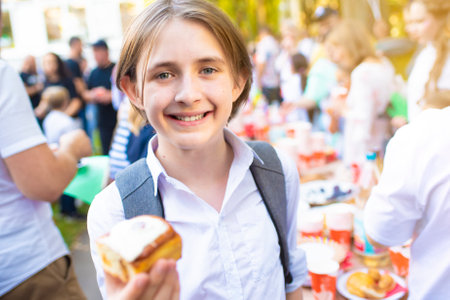 A boy in a school uniform and with a backpack in his hands holds a bun while standing in front of a festive table at a tea party at schoolの写真素材