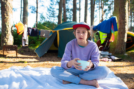An 11 year old boy or teenager has breakfast with cereal and milk, sitting on a blanket in front of a tent in the forest, spending summer holidays in a tent camp.の写真素材
