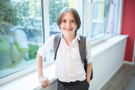 Portrait of a schoolboy with backpack standing by window at schoolの写真素材