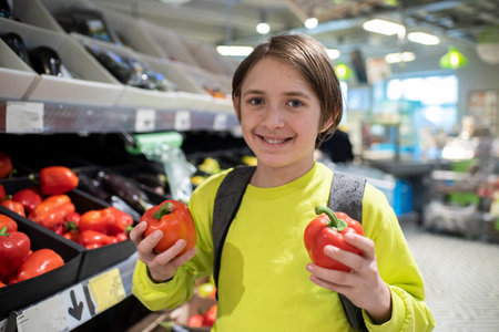 Smiling 11 year old boy with backpack holding red pepper in vegetable section of grocery store.の写真素材