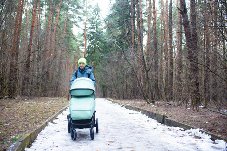 A 12 year old boy walks with a green stroller with his sleeping brother inside, in a forest or park in late winter or early spring.の写真素材