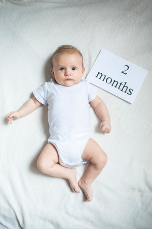 A baby boy in a white bodysuit lies on a white bed. The child is 61 days old. To the right of the...
