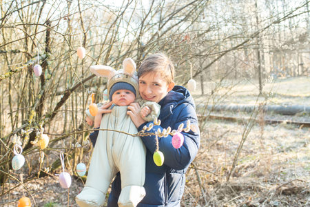 A teenage boy holds his baby brother with rabbit ears on his head, the brothers stand near a tree...