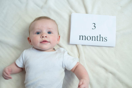 A baby boy in a white bodysuit lies on a white bed. The child is 91 days old. To the right of the child is a piece of paper with the inscription 3 months.の写真素材