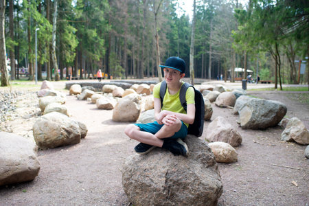 A 12-year-old boy sits on a large rock in a spring park. The teenager walks near a cascade of ponds in Borovsky Park in Moscow.の写真素材