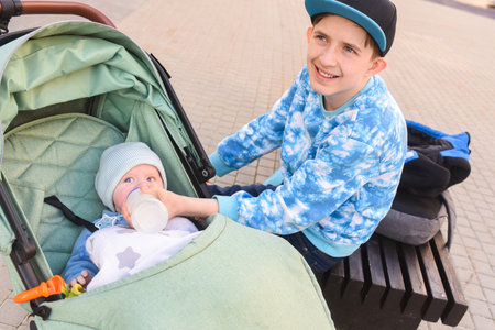 12 year old boy feeding his baby brother with milk formula in a bottle. teenager holding baby bottle, sitting on bench near green stroller with little boy. outsideの写真素材