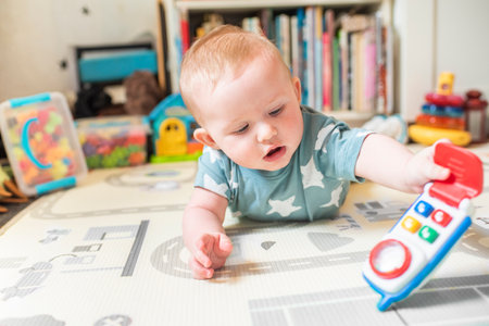 Baby boy lying in blue t-shirt with white stars lying on stomach on mat playing with toy mobile rattle at home. Child is 5 months old.の写真素材