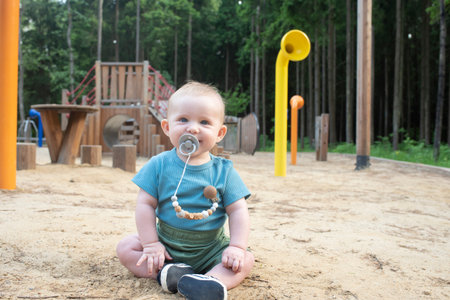 Happy baby with gray pacifier sitting on sand at playground in park in summer.の写真素材