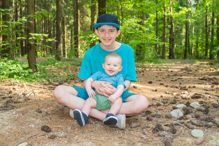 Teenage boy with his younger baby brother outdoors, siblings sitting on the ground in the forest, fir-tree cones lying around.の写真素材