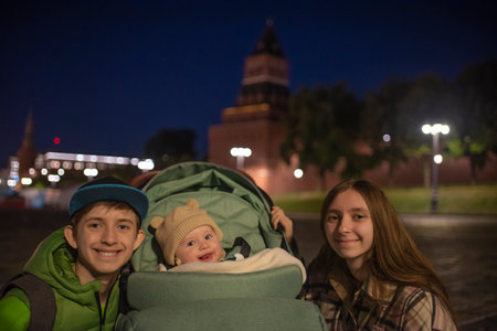 Brothers and sister are on red square near kremlin wall in center Moscow at night. boy and girl teenagers and younger baby brother in a green stroller in historical center.の写真素材