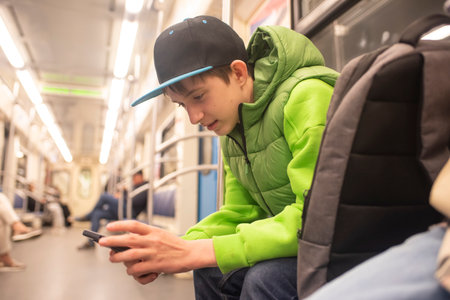 a 12 year old boy in a cap and green jacket playing a game on a mobile phone while sitting in a subway car.の写真素材