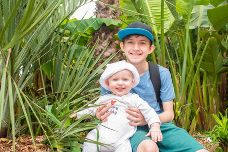 smiling teenage boy with his baby brother sitting among palm treesの写真素材