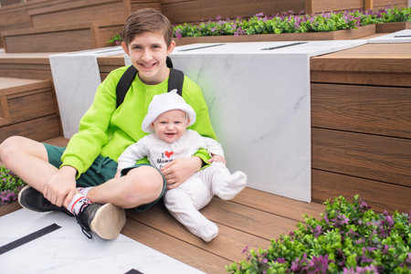 Happy 12 year old boy hugging his baby brother, children sitting on bench in park.の写真素材