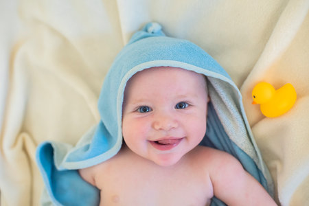 Smiling baby boy in blue towel with hoodie lying after shower on bed with yellow duck for bath.の写真素材