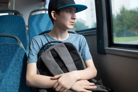 A 12 year old boy in a cap, with a backpack, sits by the window on a bus. A teenager rides alone on public transport in the summer.の写真素材