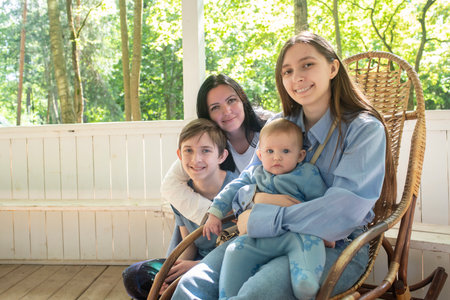 mother with three children pose for photo. brother and sister teenagers, baby brother and mother sit in white gazebo in summer outdoors.の写真素材
