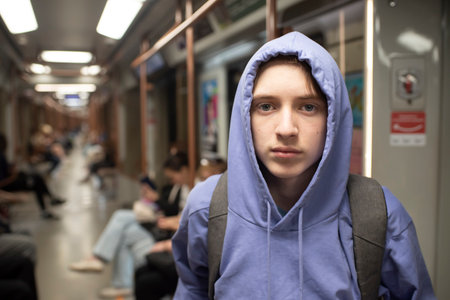 A serious teenage boy in a purple hoodie and with a backpack stands in a subway car.の写真素材