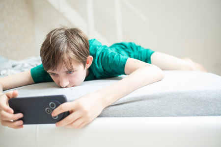 The boy is lying on the bed, holding a mobile phone in his hands, looking at the screen.の写真素材