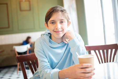 Portrait of a 12 year old boy in a blue hoodie, sitting in a cafe at a table with a paper cup of coffee.の写真素材