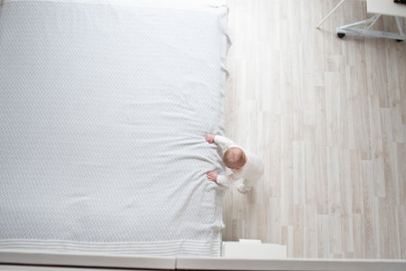 A small child, a baby in white clothes, holds on to a large parental bed and stands in a white room....
