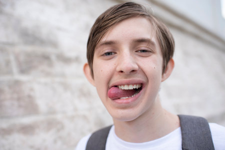 Portrait of 12 year old smiling joyful boy is showing the tongue. Child is in white t-shirt and with backpack on street in summer.の写真素材