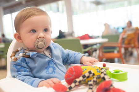 A baby boy in blue clothes sits on a high chair in a cafe, a colorful soft toy lies in front of the...
